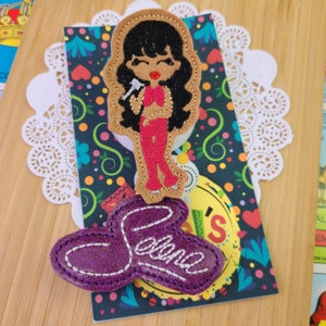 May include: A felt applique featuring a cartoon image of a singer in a red outfit, holding a microphone. Below is a purple applique with the name "Selena" in white stitching. The appliques are on a patterned card.