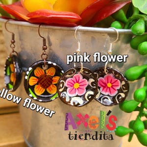 K&ouml;nnte beinhalten: Handbemalte, runde Ohrringe mit floralen Designs. Ein Paar zeigt eine gelbe Blume, ein anderes eine rosa Blume und ein drittes mit einer Mischung aus Farben. Die Ohrringe werden in einem Metalleimer mit Blumen und Gr&uuml;nzeug ausgestellt.