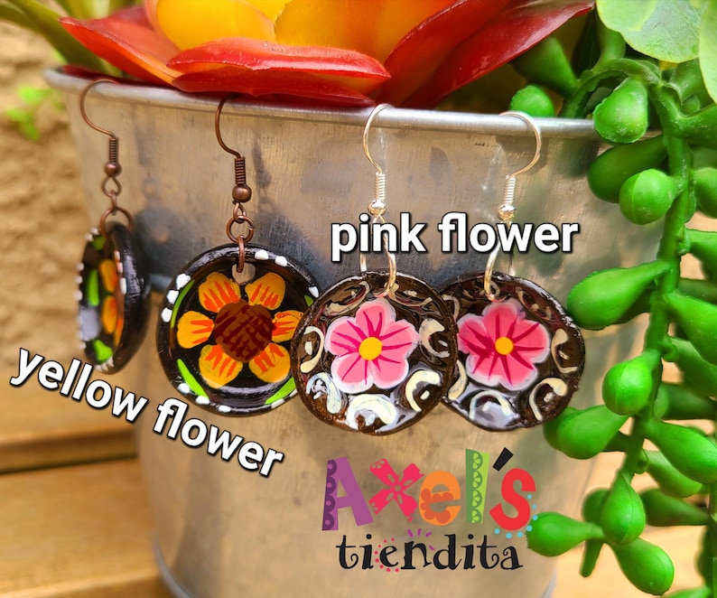 K&ouml;nnte beinhalten: Handbemalte, runde Ohrringe mit floralen Designs. Ein Paar zeigt eine gelbe Blume, ein anderes eine rosa Blume und ein drittes mit einer Mischung aus Farben. Die Ohrringe werden in einem Metalleimer mit Blumen und Gr&uuml;nzeug ausgestellt.
