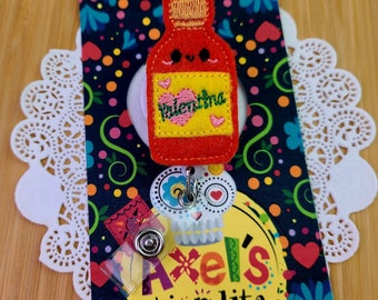 May include: A red felt badge reel in the shape of a hot sauce bottle, labelled "Valentina". The badge reel is attached to a colourful card with the text "Axel's tiendita". A white doily is in the background.