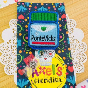 May include: A blue and green embroidered badge shaped like a jar of ointment, with the text "PonteVicks". The badge is attached to a retractable badge reel with a Day of the Dead skull design and the text "Axel's tiendita".