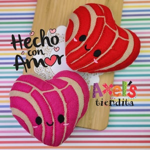 May include: Two heart-shaped plush toys, one red and one pink, resembling concha pastries, with smiling faces. The toys rest on a wooden board with the text "Hecho con Amor" and "Axel's tiendita".