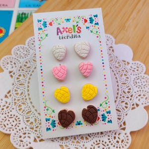 May include: A set of six colorful concha-shaped stud earrings in white, pink, yellow, and brown. The earrings are displayed on a white card with the text "Axel's tiendita" and decorative floral elements.