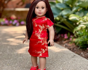 Puede incluir: Una muñeca con un vestido rojo con bordados florales dorados y zapatos rojos. La muñeca tiene el pelo largo y castaño y los ojos marrones.