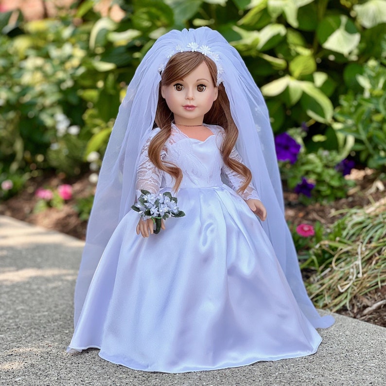 May include: A white satin bridal gown with lace sleeves and a veil. The doll is holding a small bouquet of white flowers.