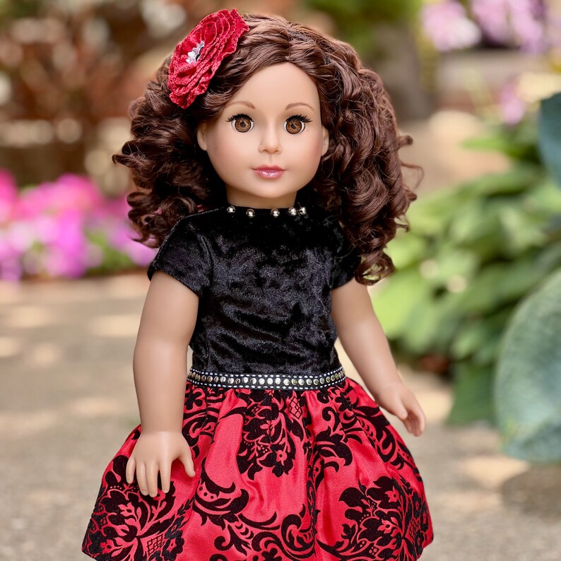 May include: A doll with brown curly hair and brown eyes, wearing a red flower hair accessory. The doll is dressed in a black velvet short-sleeved top with silver studs at the neckline and a red skirt featuring a black damask pattern.