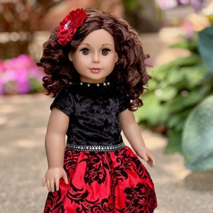 May include: A doll with brown curly hair and brown eyes, wearing a red flower hair accessory. The doll is dressed in a black velvet short-sleeved top with silver studs at the neckline and a red skirt featuring a black damask floral pattern.