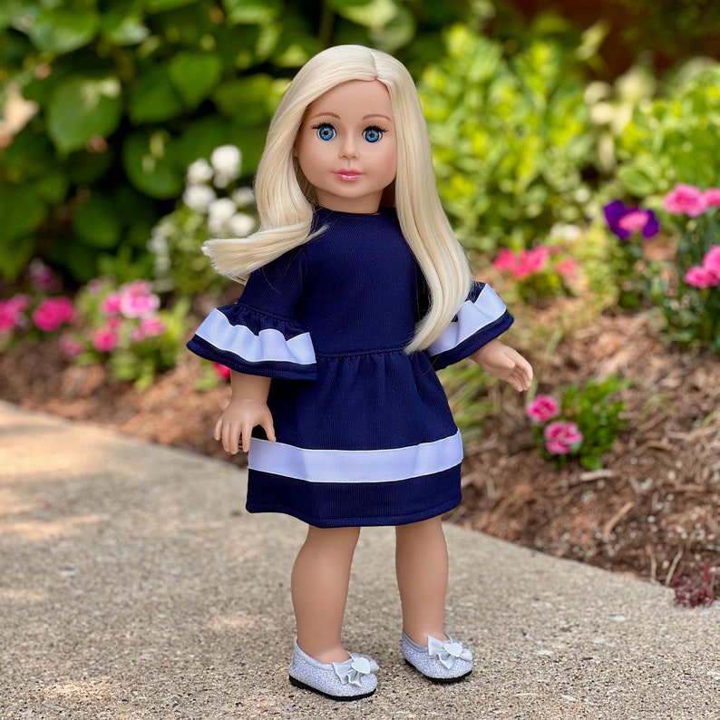 K&ouml;nnte beinhalten: Eine Puppe mit blonden Haaren, die ein marineblaues Kleid mit wei&szlig;en Verzierungen und wei&szlig;en Schuhen tr&auml;gt.