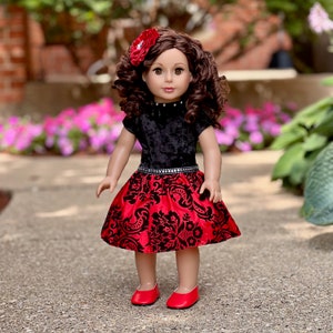 May include: A doll with curly brown hair styled with a red flower accessory. The doll wears a black velvet top with a high neckline and a red skirt featuring a black damask pattern. A silver studded belt accents the waist, and red shoes.