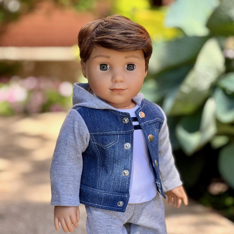 May include: A doll with brown hair and grey eyes wearing a blue denim jacket with grey jersey sleeves and an attached hood. The jacket features silver-toned metal buttons. Underneath, the doll wears a white shirt with thin horizontal stripes.