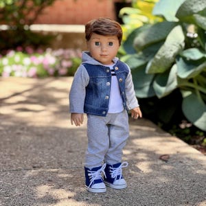 May include: A doll with brown hair and brown eyes wearing a blue denim vest with a gray hood and sleeves over a white shirt with thin black stripes. The doll is dressed in gray sweatpants and blue canvas sneakers with white laces and soles.