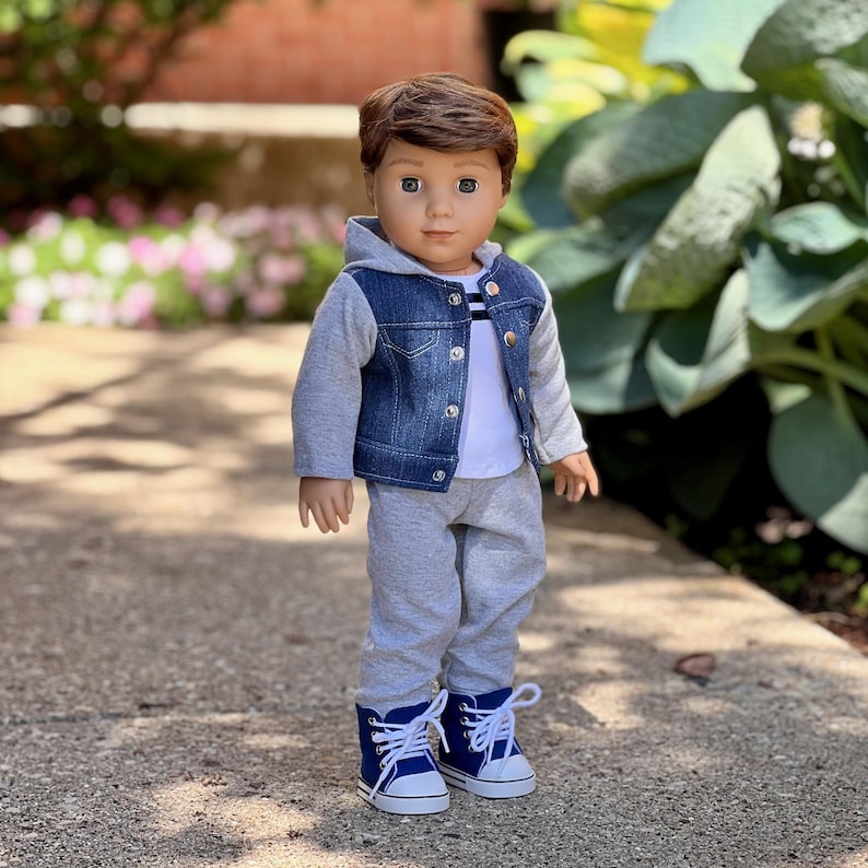 May include: A doll with brown hair and brown eyes wearing a blue denim vest with a gray hood and sleeves over a white shirt with thin black stripes. The doll is dressed in gray sweatpants and blue canvas sneakers with white laces and soles.