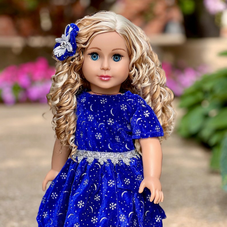 K&ouml;nnte beinhalten: Eine Puppe mit blonden, lockigen Haaren, die ein blaues Samtkleid mit silbernen Glitzer-Schneeflocken und Monden tr&auml;gt. Das Kleid hat einen silbernen Glitzerg&uuml;rtel und die Puppe tr&auml;gt eine silberne Glitzer-Haarblende.