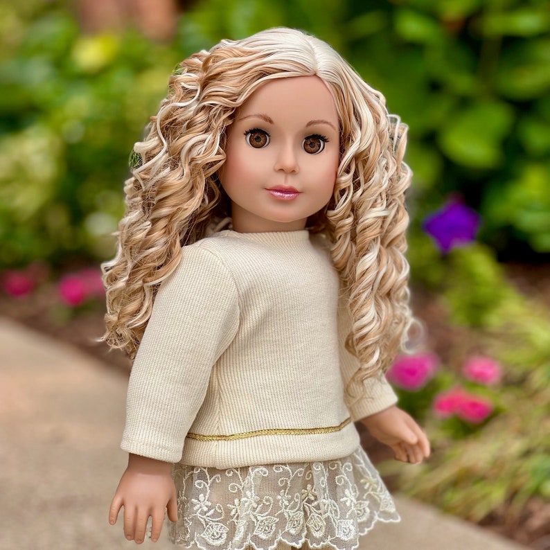 May include: A doll with long, curly blonde hair and brown eyes. She is wearing a cream-colored sweater with a gold trim and a lace skirt.