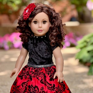 May include: A doll with long, curly brown hair and brown eyes, wearing a red fabric flower hair accessory. The doll is dressed in a black velvet short-sleeved top with silver studs at the neckline and a red skirt featuring a black floral pattern.