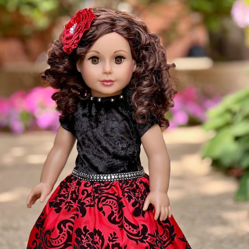 May include: A doll with long, curly brown hair and brown eyes, wearing a red fabric flower hair accessory. The doll is dressed in a black velvet short-sleeved top with silver studs at the neckline and a red skirt featuring a black floral pattern.