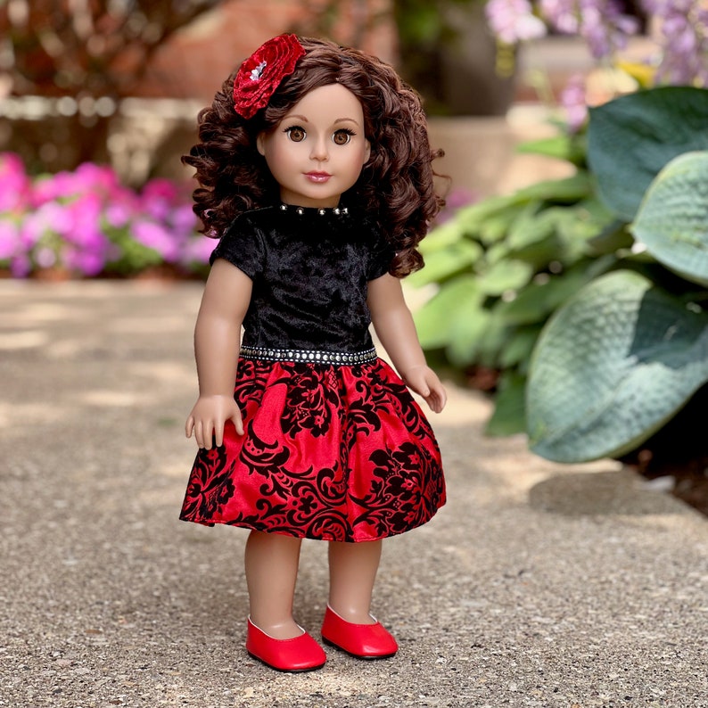 May include: A doll with curly brown hair, brown eyes, and a red flower hair accessory. The doll wears a black velvet short-sleeved top with a silver-studded neckline and a red skirt featuring a black damask pattern, paired with red shoes.