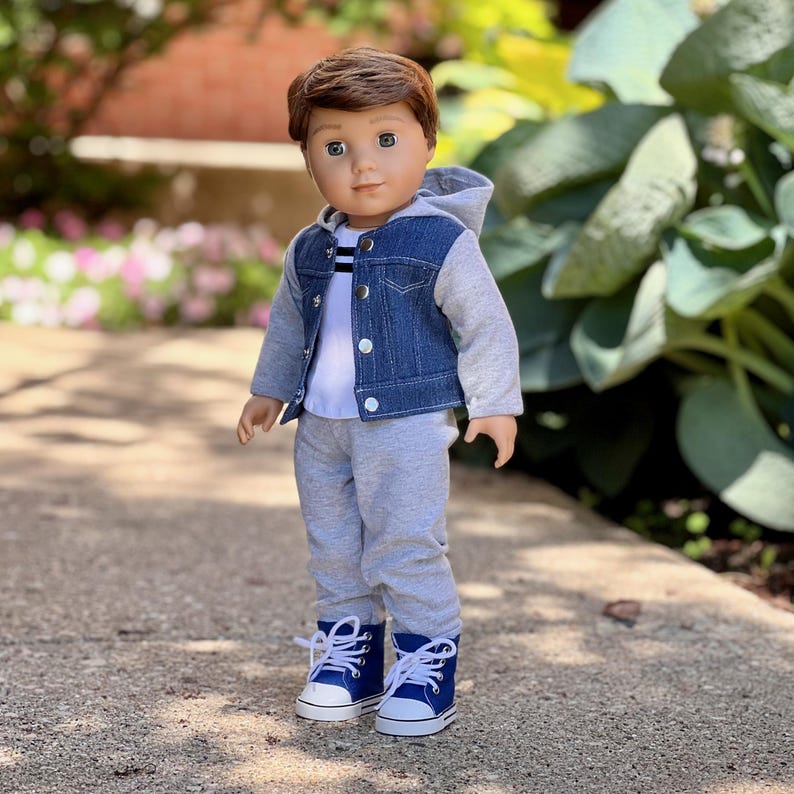 May include: A doll with brown hair and blue eyes wearing a white t-shirt with black stripes, a blue denim vest with a gray hood and sleeves, gray sweatpants, and blue high-top sneakers with white laces and white rubber toe caps.