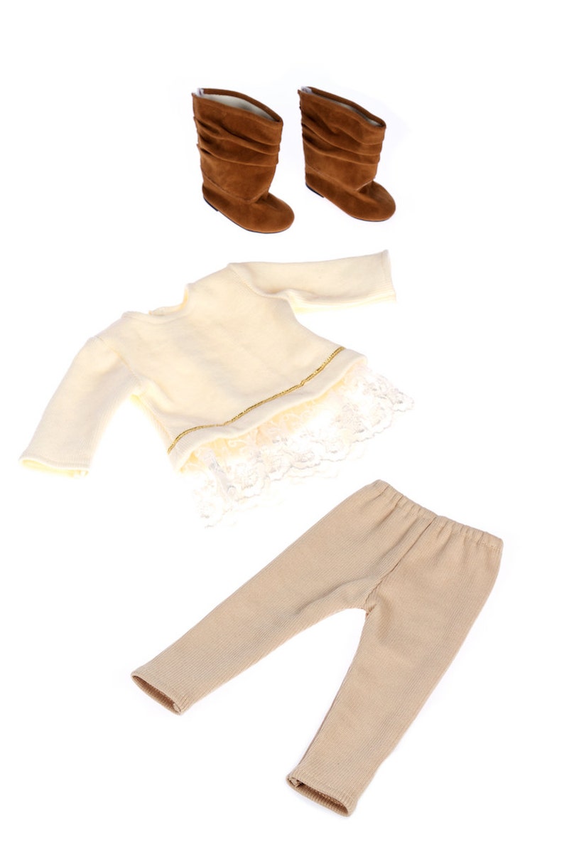 May include: A three-piece outfit for a doll, including a cream-colored sweater with lace trim, beige leggings, and brown suede boots.