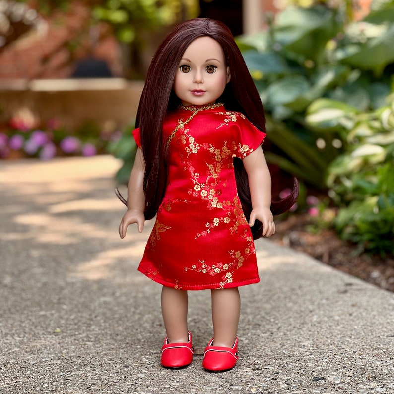 May include: A doll wearing a red and gold Chinese-style dress with floral patterns. The doll has long brown hair and brown eyes. She is wearing red shoes.