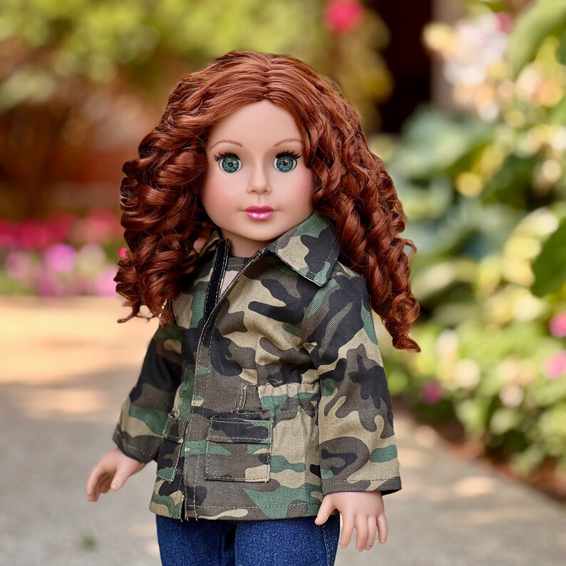 May include: A doll with long, curly auburn hair and green eyes. The doll is wearing a camouflage print jacket with a collar, long sleeves, and two front pockets, paired with dark blue denim trousers. The doll has a neutral expression and pink lips.