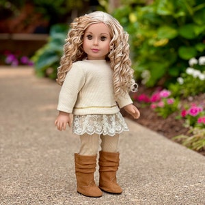 May include: A doll wearing a cream-colored sweater with a gold trim, a lace skirt, beige leggings, and brown boots. The doll has long, curly blonde hair and blue eyes.