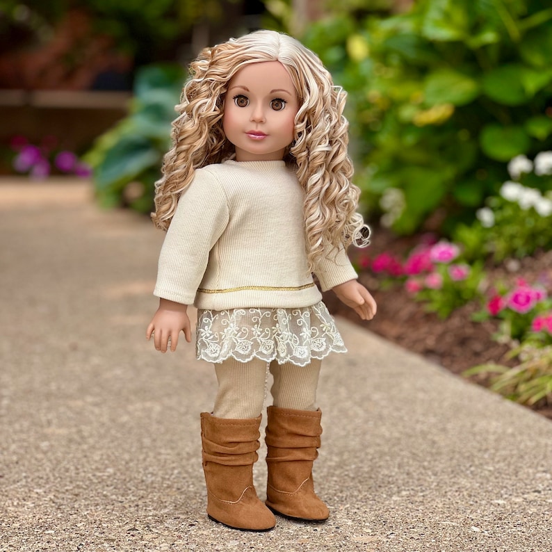 May include: A doll wearing a cream-colored sweater with a gold trim, a lace skirt, beige leggings, and brown boots. The doll has long, curly blonde hair and blue eyes.