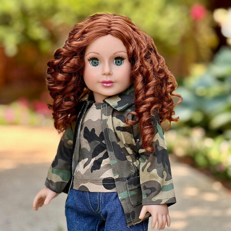 May include: A doll with long, curly auburn hair and green eyes, wearing a camouflage print jacket over a matching camouflage t-shirt and dark blue denim jeans. The doll has a fair complexion and pink lips, with the jacket featuring a collar.