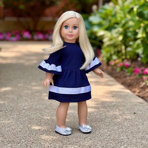 K&ouml;nnte beinhalten: Eine Puppe, die ein marineblaues Kleid mit wei&szlig;en Verzierungen und wei&szlig;en Schuhen tr&auml;gt. Das Kleid hat Glocken&auml;rmel und ein wei&szlig;es Band um die Taille.