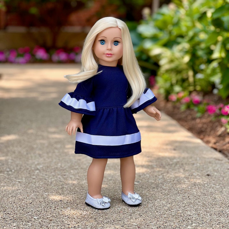 K&ouml;nnte beinhalten: Eine Puppe, die ein marineblaues Kleid mit wei&szlig;en Verzierungen und wei&szlig;en Schuhen tr&auml;gt. Das Kleid hat Glocken&auml;rmel und ein wei&szlig;es Band um die Taille.
