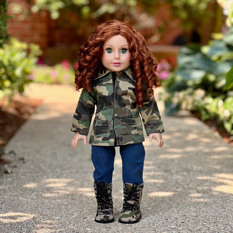 May include: A doll with long curly auburn hair and green eyes wearing a camouflage print jacket with two front pockets, dark blue denim trousers, and camouflage print lace-up boots with black laces. The doll has a neutral expression and stands upright.