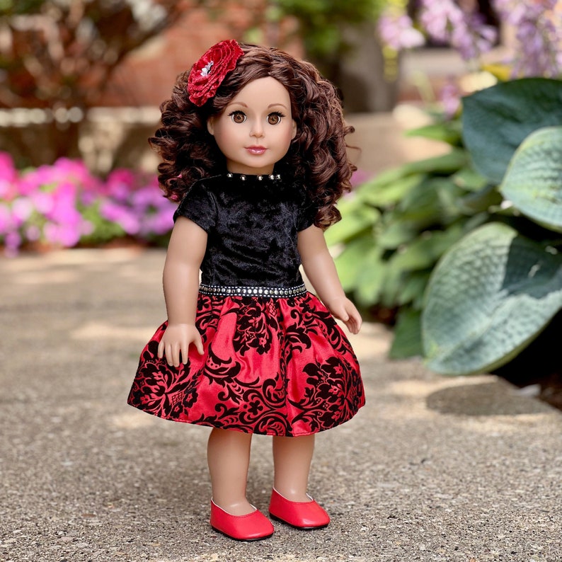 May include: A doll with brown curly hair and a red flower hair accessory. The doll wears a black velvet short-sleeved top with a silver-studded neckline and a red skirt featuring a black damask pattern and a silver-studded waistband, plus red shoes.