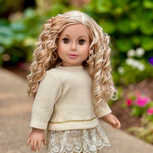 May include: A doll with long, curly blonde hair wearing a cream-colored sweater with a gold trim and a white lace skirt.