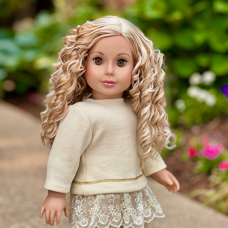 May include: A doll with long, curly blonde hair wearing a cream-colored sweater with a gold trim and a white lace skirt.