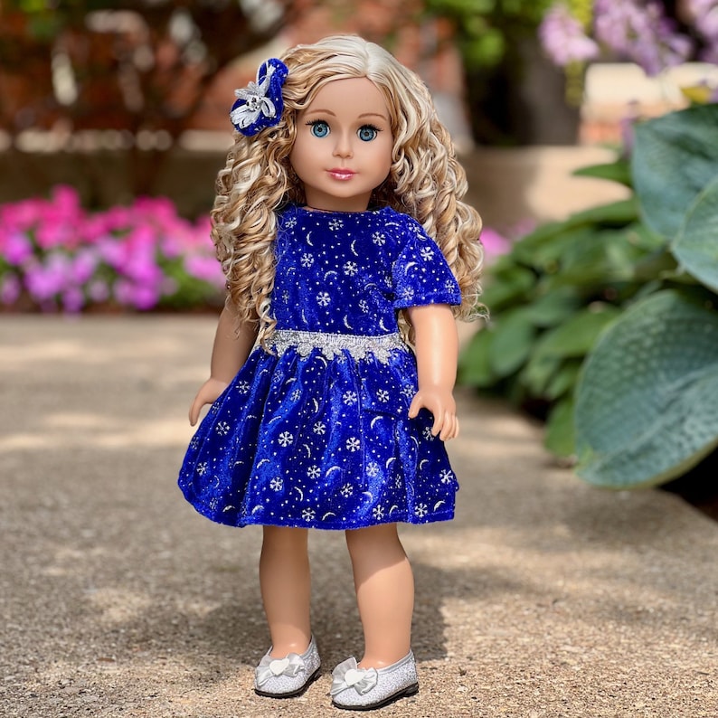 K&ouml;nnte beinhalten: Eine Puppe tr&auml;gt ein blaues Samtkleid mit einem silbernen Schneeflocken- und Halbmondmuster. Das Kleid hat einen silbernen Besatz um die Taille. Die Puppe hat blonde lockige Haare und blaue Augen. Sie tr&auml;gt silberne Schuhe.