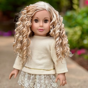 May include: A doll with long, curly blonde and white hair wearing a cream-colored sweater with a gold trim and a lace skirt.