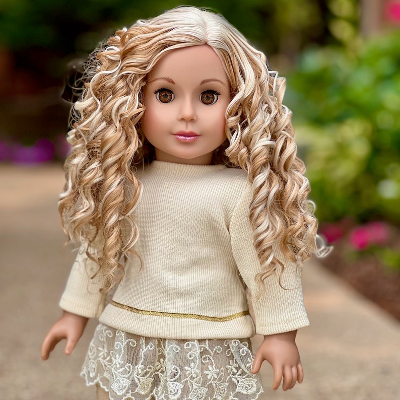 May include: A doll with long, curly blonde and white hair wearing a cream-colored sweater with a gold trim and a lace skirt.