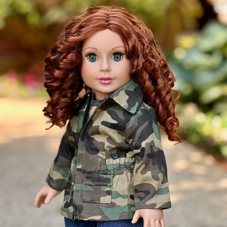May include: A doll with long, curly auburn hair and green eyes, wearing a camouflage print jacket with a collar, long sleeves, and two front pockets. The jacket features a cinched waist design and is worn over a dark blue garment.