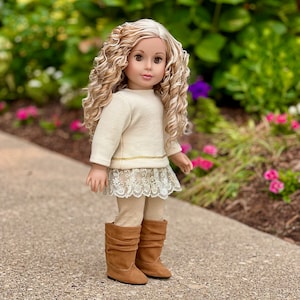 May include: A doll with long blonde curly hair wearing a cream colored sweater, a lace skirt, beige leggings, and brown boots.