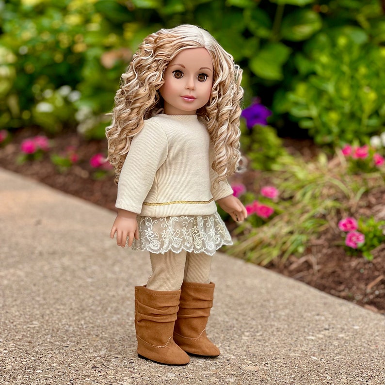 May include: A doll with long blonde curly hair wearing a cream colored sweater, a lace skirt, beige leggings, and brown boots.