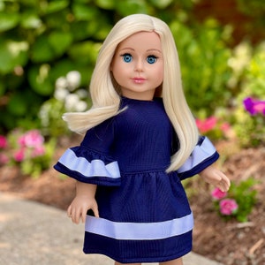 K&ouml;nnte beinhalten: Eine Puppe in einem dunkelblauen Kleid mit wei&szlig;en Verzierungen und wei&szlig;en, gebauschten &Auml;rmeln. Die Puppe hat langes blondes Haar und blaue Augen.