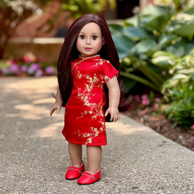 May include: A doll wearing a red dress with gold floral embroidery and red shoes. The doll has long brown hair and brown eyes.