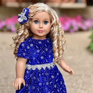 K&ouml;nnte beinhalten: Eine Puppe mit blonden, lockigen Haaren, die ein blaues Samtkleid mit silbernen Schneeflocken und Monden tr&auml;gt. Das Kleid hat einen silbernen Spitzenbesatz. Die Puppe tr&auml;gt eine silberne und wei&szlig;e Schleife im Haar.