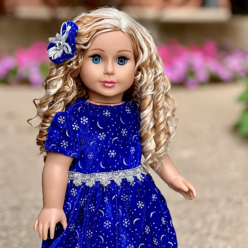 K&ouml;nnte beinhalten: Eine Puppe mit blonden, lockigen Haaren, die ein blaues Samtkleid mit silbernen Schneeflocken und Monden tr&auml;gt. Das Kleid hat einen silbernen Spitzenbesatz. Die Puppe tr&auml;gt eine silberne und wei&szlig;e Schleife im Haar.