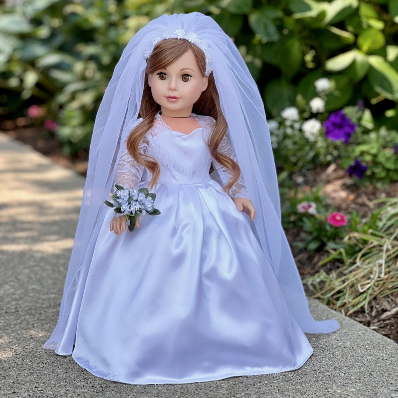 May include: A white satin wedding dress with lace sleeves and a long veil. The dress has a sweetheart neckline and a fitted bodice. The doll is holding a small bouquet of white flowers.