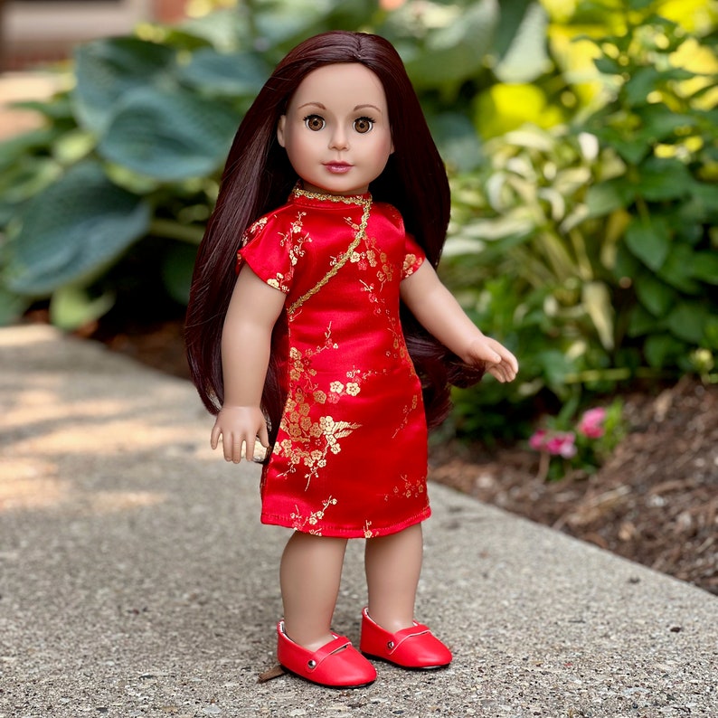 May include: A doll wearing a red and gold Chinese-style dress with floral patterns. The doll has long brown hair and brown eyes.