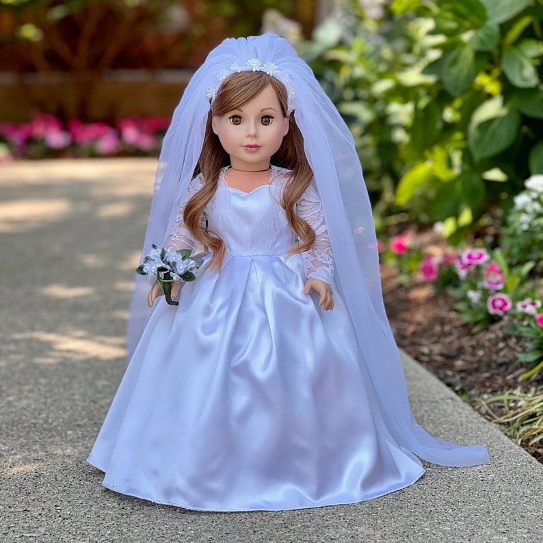 May include: A white satin wedding dress with lace sleeves and a tulle veil for a doll. The dress has a sweetheart neckline and a full skirt. The doll is holding a small bouquet of white flowers.