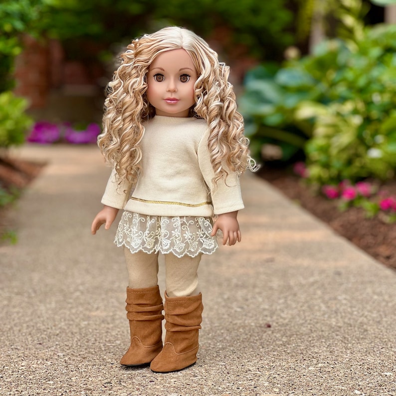 May include: A doll wearing a cream-colored sweater with a lace trim and brown boots. The doll has long blonde curly hair and brown eyes.