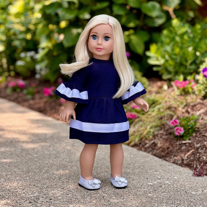 K&ouml;nnte beinhalten: Eine Puppe in einem dunkelblauen Kleid mit wei&szlig;en Verzierungen und wei&szlig;en Schuhen. Die Puppe hat blonde Haare und blaue Augen.
