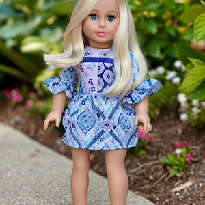 May include: A blonde doll wearing a blue and white patterned dress with silver shoes. The doll has blue eyes and is standing on a gray sidewalk.